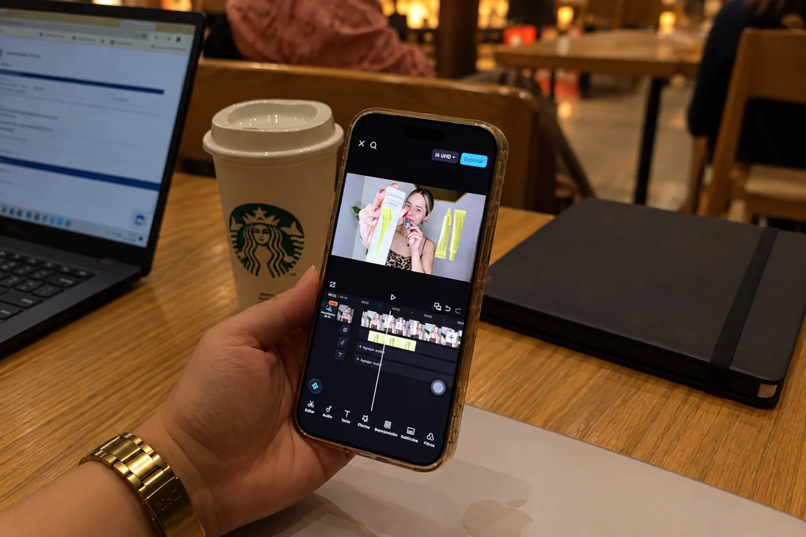 Celular mostrando una app de edición de video con un clip en reproducción, laptop y café de fondo en una cafetería