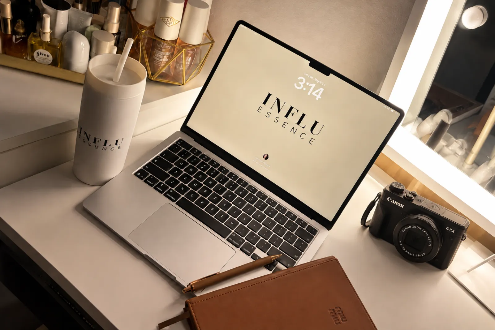 Escritorio con laptop mostrando la pantalla de inicio de Influessence Academy, cámara, libreta de notas y productos de belleza alrededor