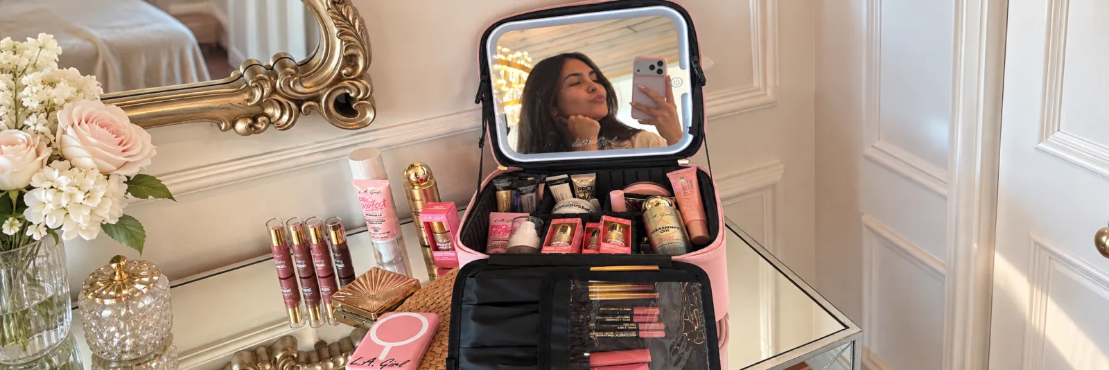 Mujer tomándose una selfie frente a un espejo con luces, rodeada de productos de maquillaje organizados en un estuche