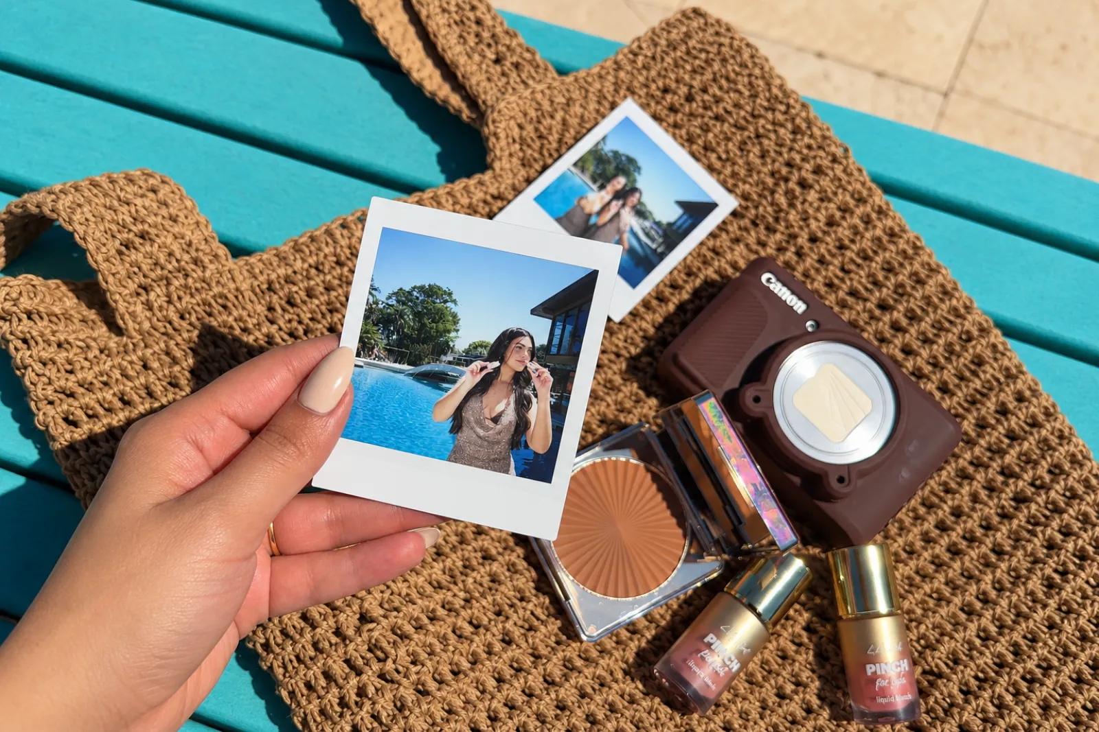 Mano sosteniendo fotos instantáneas junto a una cámara y productos de maquillaje sobre un bolso tejido, ambiente de playa